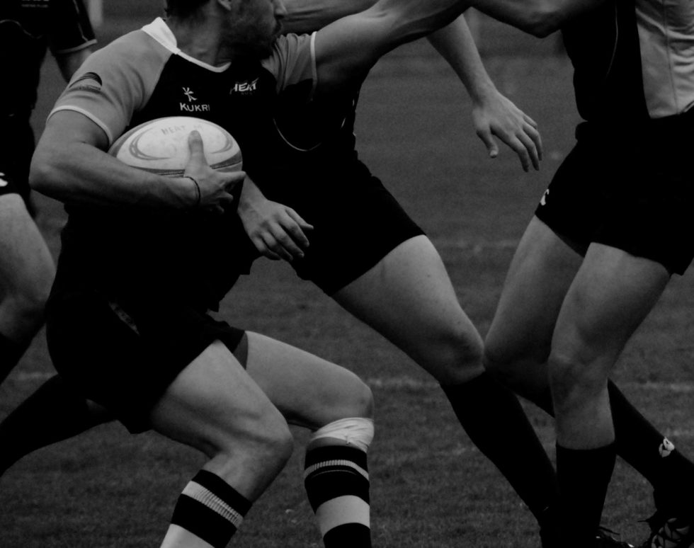 A rugby player holding a ball tries to evade defenders, one of whom is reaching out an arm to block him. The image, in black and white, highlights the intensity and movement&mdash;much like bespoke sports kits bring unique energy to every match.