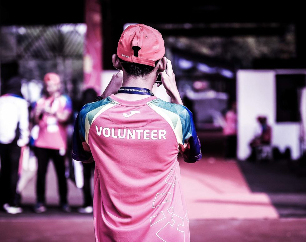 Eine Person mit roter Mütze und einem Hemd mit der Aufschrift VOLUNTEER steht im Freien und macht möglicherweise Fotos von maßgeschneiderten Fußballtrikots. Andere Personen und ein Gebäude sind unscharf im Hintergrund zu erkennen.