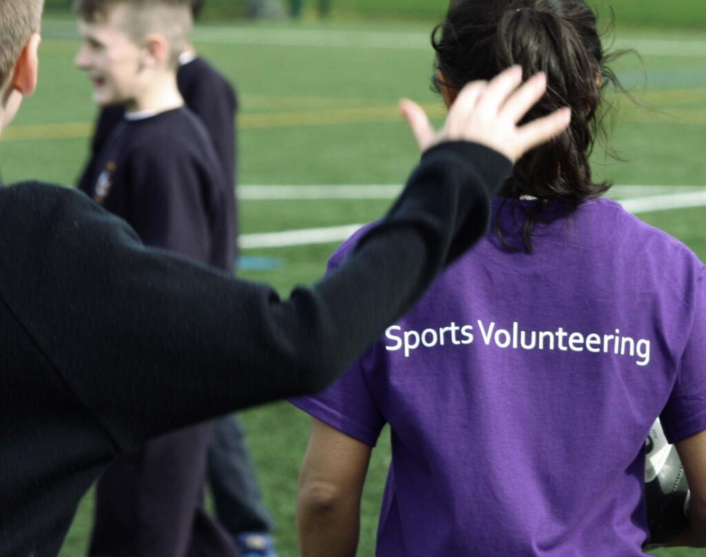 Une personne portant un kit de sport violet personnalisé tient un ballon sur un terrain de sport, tandis qu'une autre personne tend la main vers elle. Des enfants flous sont visibles à l'arrière-plan.