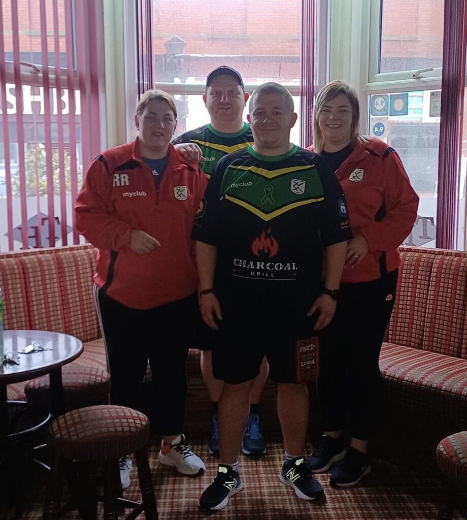 Four adults stand together in a pub with chequered seating and large windows. Three wear red jackets with club logos, while one in front sports a custom football shirt designed by a leading kit designer, featuring "Charcoal Grill" on the chest.
