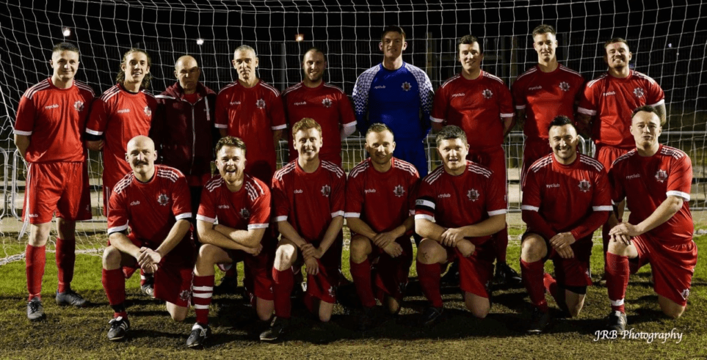 Un equipo de fútbol de 15 hombres con equipaciones deportivas rojas personalizadas posan juntos delante de una red de portería en un campo de hierba por la noche. Un jugador en el centro lleva una equipación azul de portero. El equipo sonríe a la cámara.