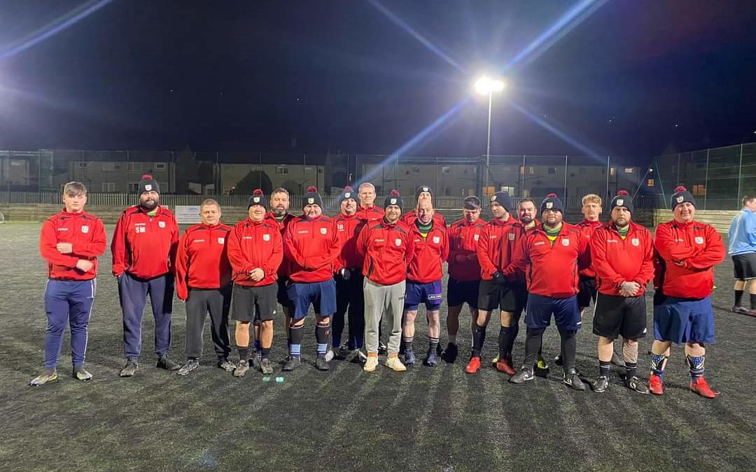 A group of men in matching red personalised football kits and black shorts stand together on an outdoor football pitch at night, posing for a team photo under bright floodlights.