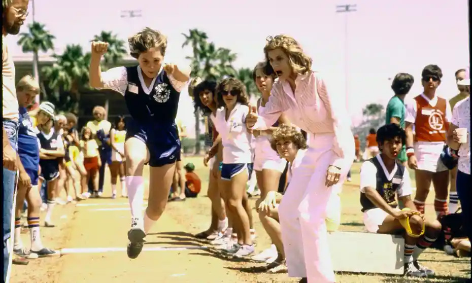 Eine junge Athletin in maßgeschneiderter Sportkleidung springt während einer Leichtathletikveranstaltung, während eine Frau in Rosa sie anfeuert. Zuschauer und andere Teilnehmer schauen unter sonnigem Himmel mit Palmen im Hintergrund zu.