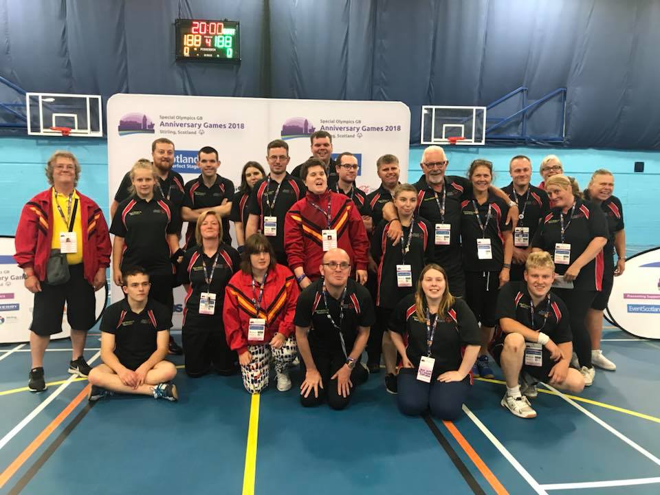 Eine große Gruppe von Athleten und Trainern in passender schwarz-roter Sportkleidung posiert gemeinsam auf einem Sportplatz und lächelt vor den Bannern für die Special Olympics Anniversary Games 2018.