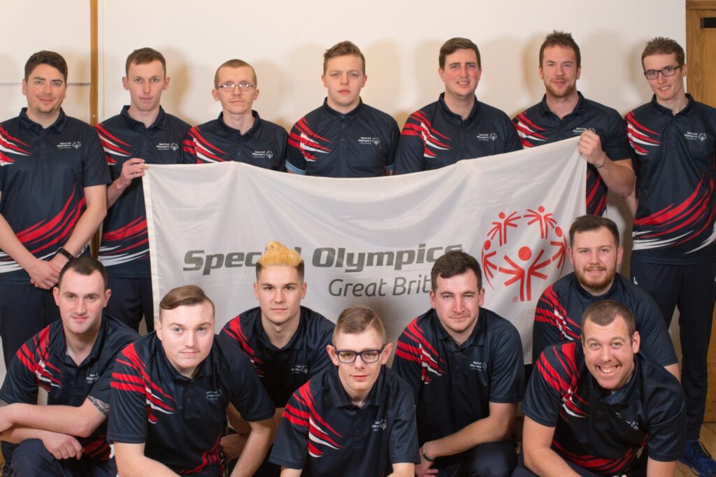 Eine Gruppe von vierzehn Männern in passender Sportkleidung mit marineblauen Hemden und roten Streifen posiert zusammen und hält ein Banner der Special Olympics Great Britain. Sie lächeln und stehen vor einer schlichten hellen Wand.