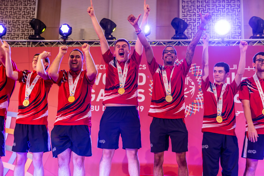 Fünf männliche Athleten in passenden Sporttrikots feiern auf der Bühne mit erhobenen Armen, lächelnd und jubelnd, jeder mit einer Goldmedaille um den Hals nach einem Sieg bei einem Sportereignis.