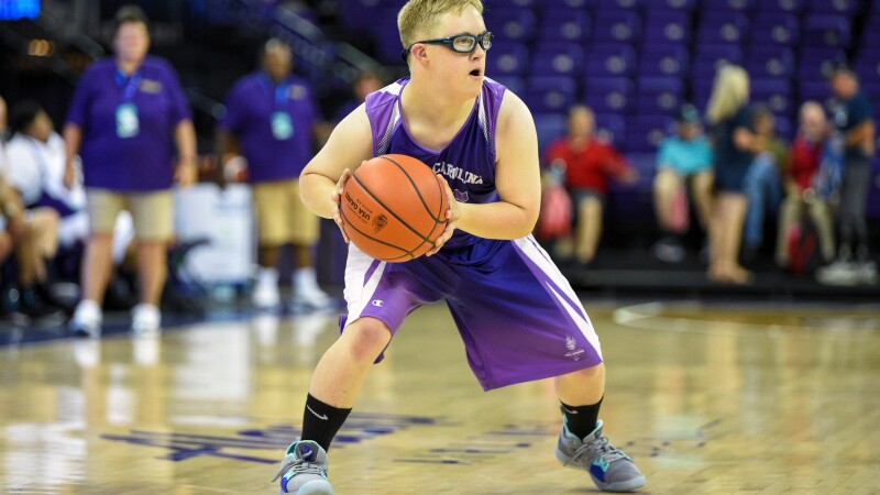 Ein junger Sportler mit Brille und lilafarbenem Sportdress hält einen Basketball auf einem Hallenplatz und bereitet sich auf einen Pass oder einen Schuss vor. Im Hintergrund sind Zuschauer und andere Spieler zu sehen.