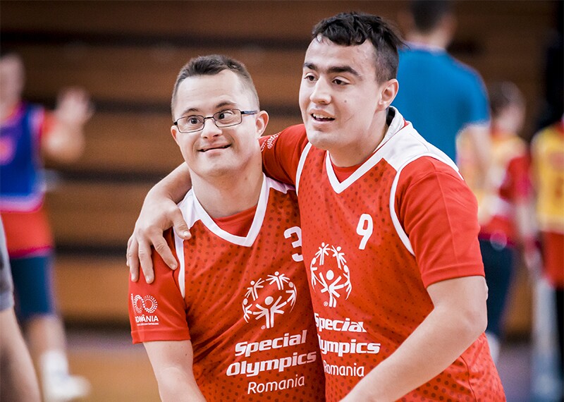 Zwei männliche Athleten in roten “Special Olympics Romania”-Trikots lächeln und feiern zusammen auf einem Basketballplatz, die Arme umeinander gelegt, und zeigen stolz ihre maßgeschneiderten Sporttrikots. Im Hintergrund sind andere Spieler und unscharfe Zuschauer zu sehen.