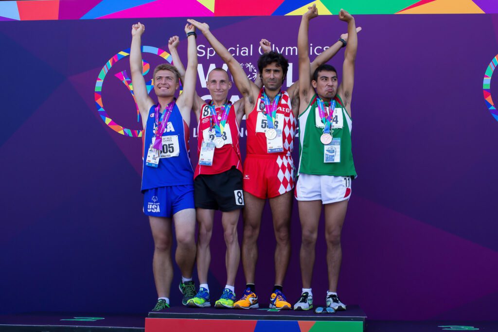 Vier männliche Athleten stehen auf einem Podium und heben jeweils einen Arm zum Sieg. Sie tragen maßgeschneiderte Sportkleidung und Medaillen um den Hals und feiern gemeinsam bei den Special Olympics World Games.