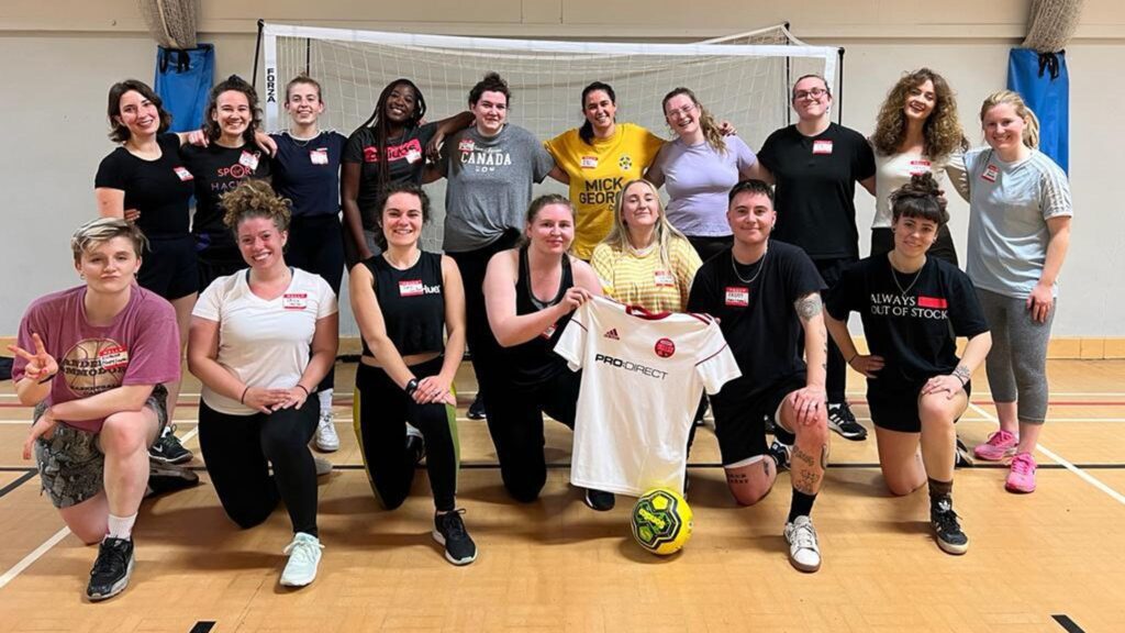 Un groupe de personnes pose et sourit dans un gymnase devant un but de football. La plupart portent des vêtements de sport, certains tiennent un ballon de football, et deux personnes à l'avant arborent fièrement leur kit de football personnalisé.