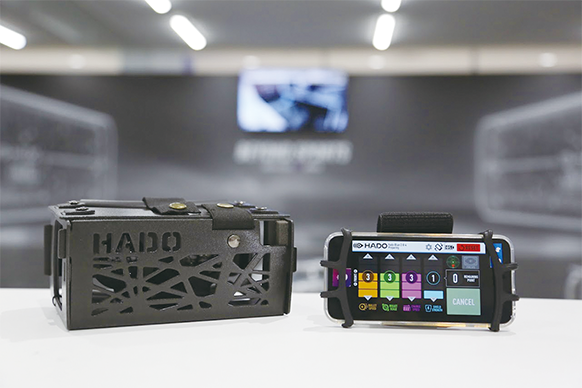 Nahaufnahme zweier HADO-Geräte auf einer weißen Fläche: ein schwarzes, rechteckiges Gehäuse mit ausgeschnittenen Mustern und HADO-Schriftzug und ein Mobiltelefon in einer Halterung mit einer bunten Schnittstelle für Designer von Sportbekleidung.