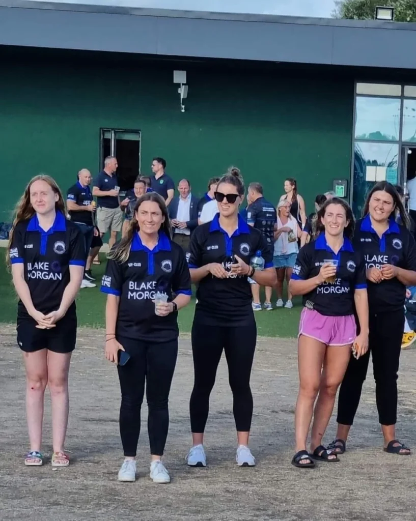 Five women in matching black and blue custom sportswear stand outdoors, smiling at the camera. A group of people and a modern building are visible in the background. Some of the women are holding drinks.