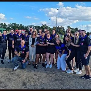 A group of people pose together outdoors on a sunny day, many wearing matching custom football shirts, standing on a pitch with green trees and a blue sky with clouds in the background.