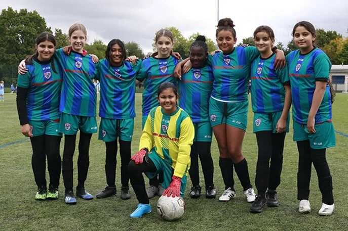 Une équipe de football féminine, vêtue de kits de football personnalisés, pose ensemble sur un terrain, en souriant à l'appareil photo. Huit joueuses se tiennent debout, les bras enlacés, et une joueuse portant un kit jaune de gardienne de but s'agenouille devant avec un ballon de football.