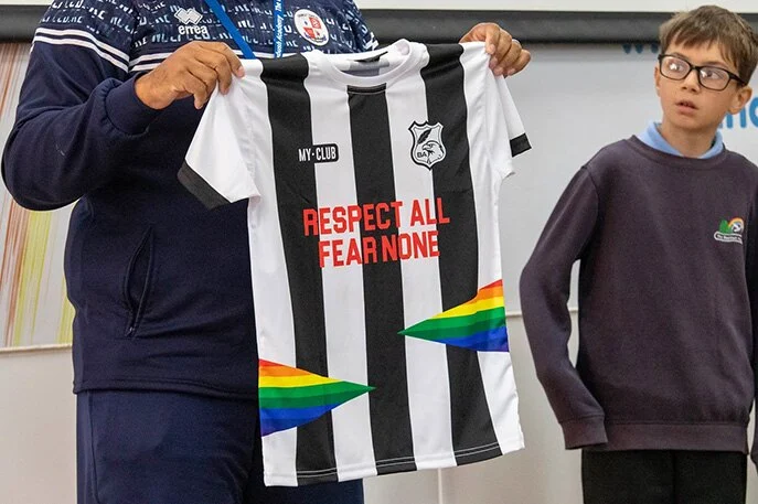 Une personne tient un maillot de sport personnalisé rayé noir et blanc avec RESPECT ALL FEAR NONE en texte rouge et aux accents arc-en-ciel, sous le regard d'un garçon portant des lunettes et un uniforme d'écolier.