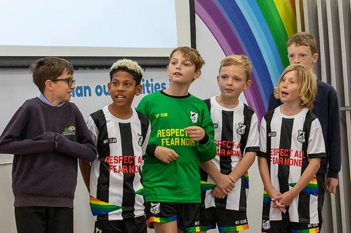 Cinq enfants vêtus de tenues de football personnalisées en noir et blanc et un autre portant un maillot vert se tiennent ensemble à l'intérieur, souriant et discutant, avec une fresque arc-en-ciel et un écran de projection à l'arrière-plan.