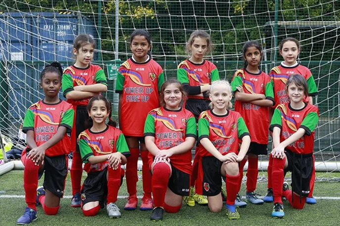 Une équipe de football féminine pose devant un but, souriante et agenouillée sur deux rangs. Elles portent des maillots de football personnalisés assortis avec l'inscription POWER UP FIRE UP sur le devant, mettant en valeur leurs vêtements de sport personnalisés sur le terrain.
