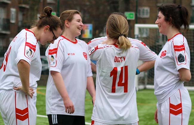 Four women in personalised football kits stand together on the pitch, smiling and chatting. One woman has her head down, while the others face each other, with buildings and a goalpost visible in the background.