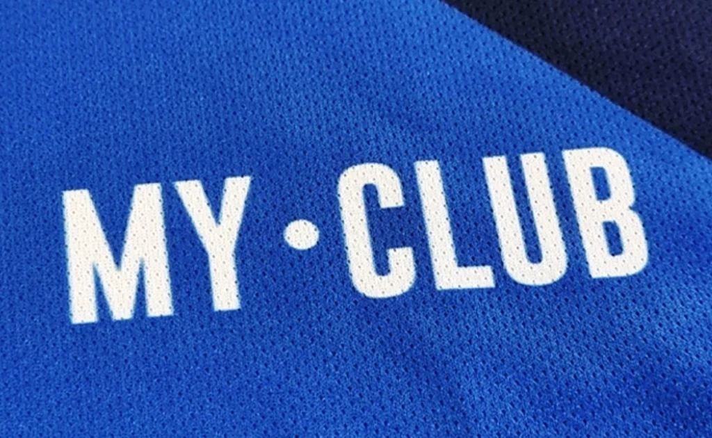 Nahaufnahme eines blauen Trikots mit der Aufschrift MY CLUB in fetten weißen Großbuchstaben, getrennt durch einen Punkt in der Mitte. Der strukturierte Stoff ist mit kleinen Perforationen versehen.