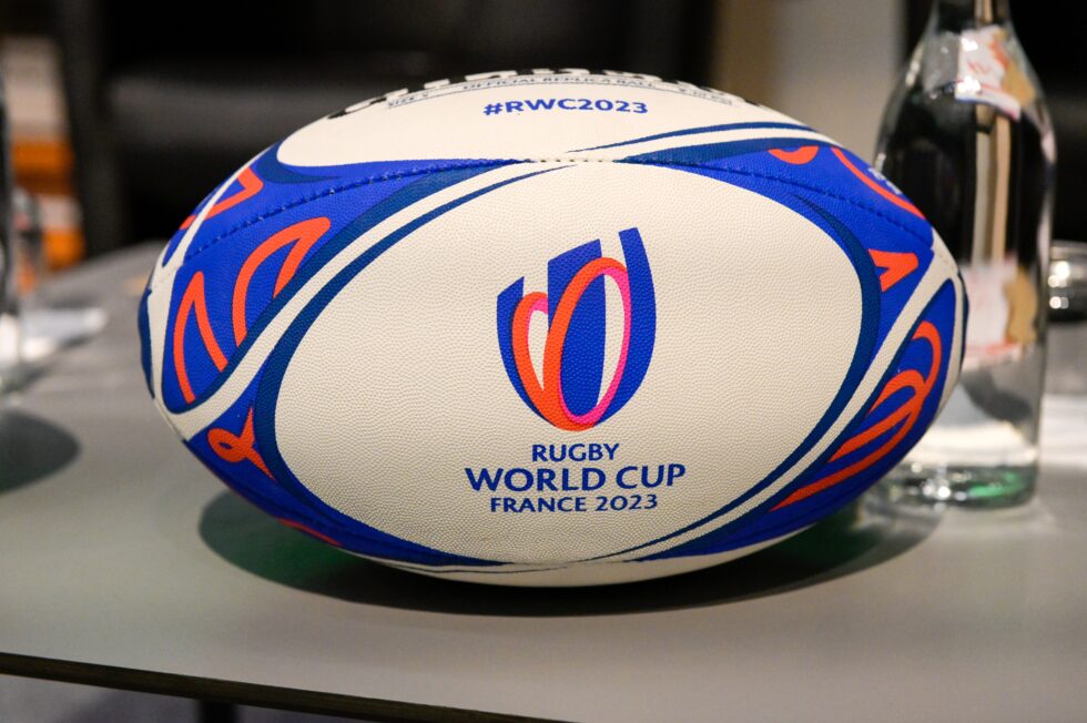 A rugby ball with blue, orange, and white patterns, featuring the Rugby World Cup France 2023 logo and hashtag #RWC2023, rests on a table next to a glass bottle&mdash;perfect inspiration for any kit designer creating custom football kits.