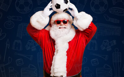 Father Christmas wearing red sunglasses and his classic red suit holds a football above his head, smiling. The blue background features faint sports-themed doodles, including footballs and custom football shirts.