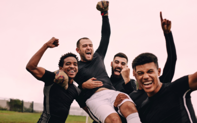 A group of excited football players in black custom football kits celebrate together, lifting a teammate into the air as they cheer and smile, showing joy and triumph on the pitch.