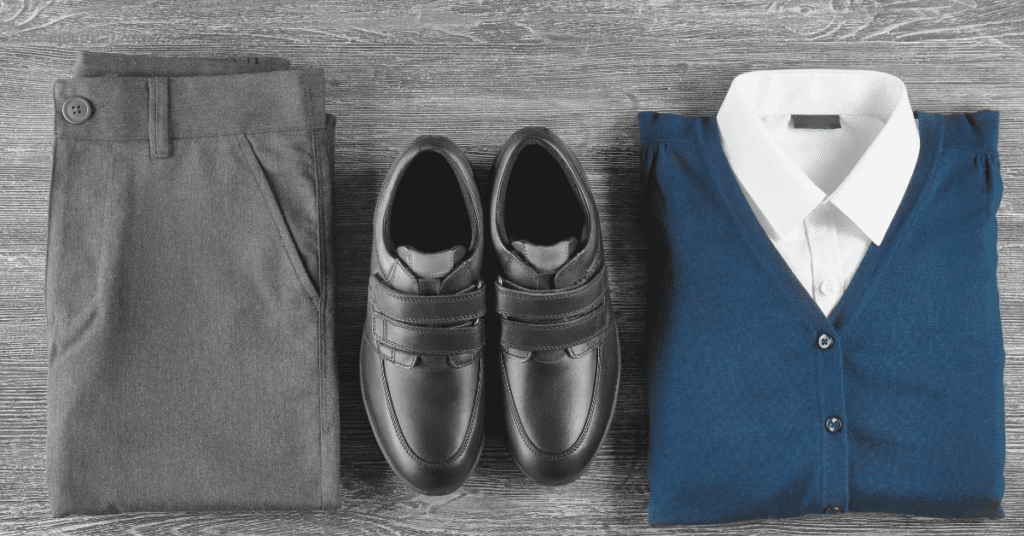 Eine graue Anzughose, schwarze Anzugschuhe und eine ordentlich gefaltete blaue Strickjacke über einem weißen Hemd mit Kragen, alles auf einer Holzfläche ausgebreitet - perfekt arrangiert wie maßgeschneiderte Sportkleidung, die auf Stil und Raffinesse zugeschnitten ist.