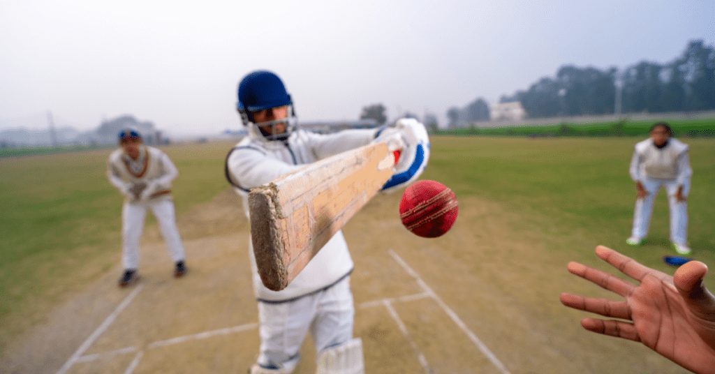 Un joueur de cricket vêtu d'une tenue et d'un casque blancs balance une batte pour frapper une balle rouge en plein vol, avec des joueurs de champ et un terrain en herbe visibles à l'arrière-plan. L'image est prise du point de vue du joueur de bowling - une inspiration parfaite pour tout concepteur de kit.
