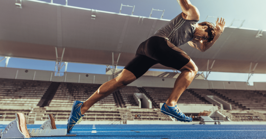 Ein männlicher Sportler in Sportkleidung sprintet auf einer blauen Laufbahn in einem leeren Stadion aus den Startblöcken. Das Sonnenlicht scheint von oben und zeigt die Geschwindigkeit und Entschlossenheit, die in jedem maßgeschneiderten Fußballtrikot steckt.