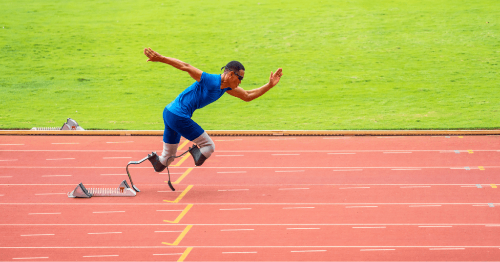 Ein männlicher Athlet mit Laufprothesen sprintet von den Startblöcken auf einer Rennbahn vorwärts. Er trägt maßgeschneiderte blaue Sportkleidung, im Hintergrund ist ein grasbewachsenes Feld zu sehen.