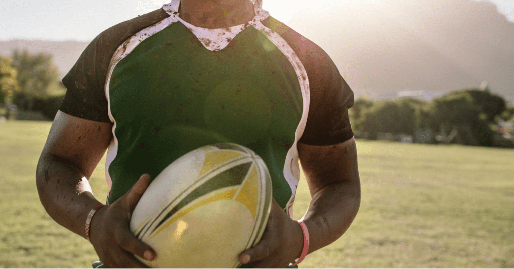 Ein Rugbyspieler in einem grün-schwarzen Trikot hält einen Rugbyball auf einem grasbewachsenen Platz, während im Hintergrund die Sonne scheint. Das Hemd des Spielers ist sichtlich mit Schlamm verschmutzt.