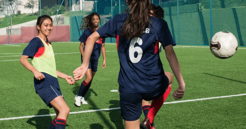 Vier Mädchen in individuellen Fußballtrikots spielen auf einem grünen Fußballplatz, wobei ein Mädchen einen Fußball kickt, während die anderen zusehen und sich auf das Spiel vorbereiten. In ihren personalisierten Fußballtrikots wirken sie während des Spiels konzentriert und aktiv.