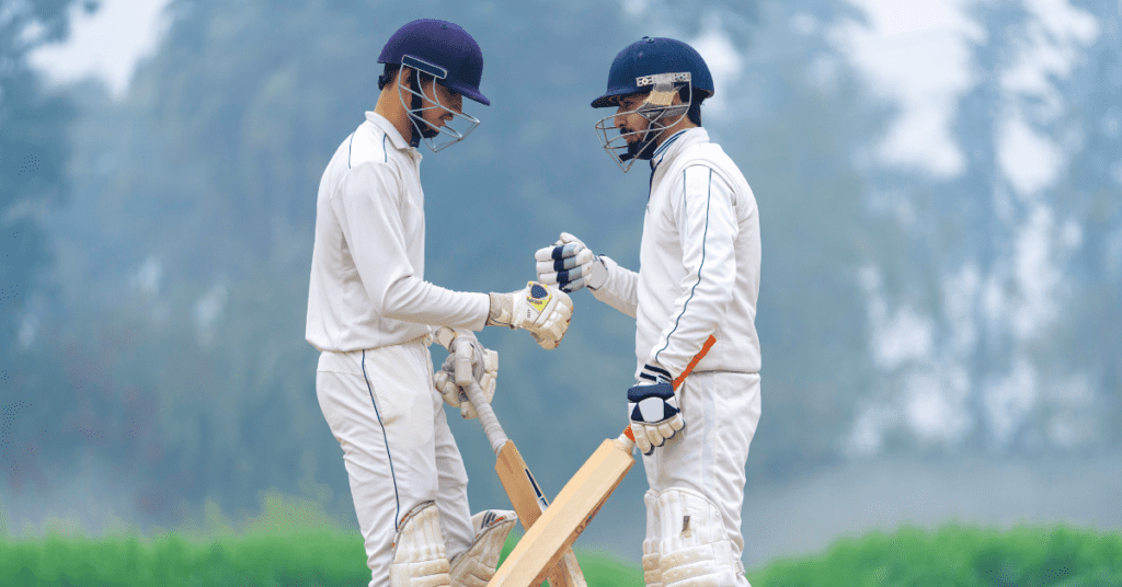 Deux joueurs de cricket vêtus d'une tenue et d'un casque blancs se frappent le poing sur le terrain, en tenant leur batte, avec de la verdure en arrière-plan - une inspiration parfaite pour tout concepteur de vêtements de sport ou de kits personnalisés.