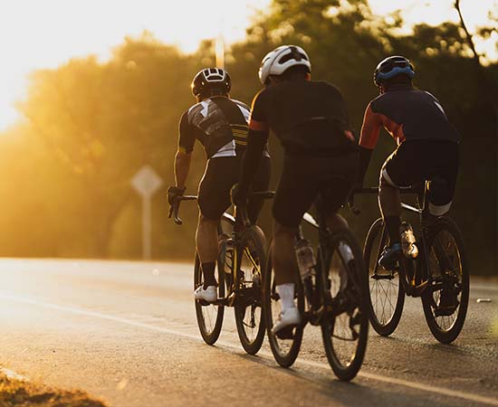 Tres ciclistas con casco y ropa deportiva personalizada circulan por una carretera asfaltada al amanecer o al atardecer, con árboles y la cálida luz del sol de fondo.
