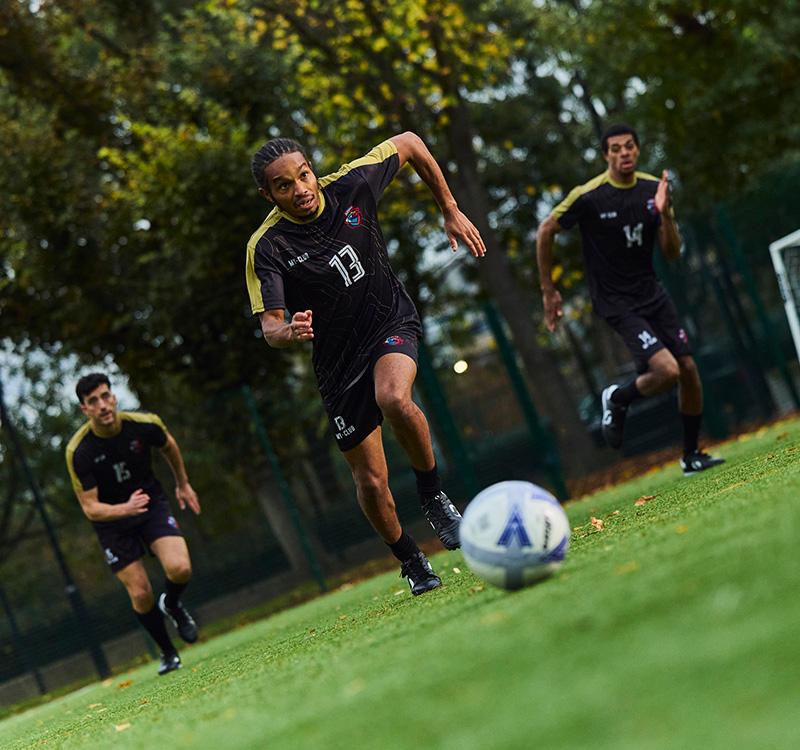 Trois joueurs de football en uniforme sombre courent sur un terrain herbeux en direction d'un ballon. Des arbres et une clôture sont visibles à l'arrière-plan.