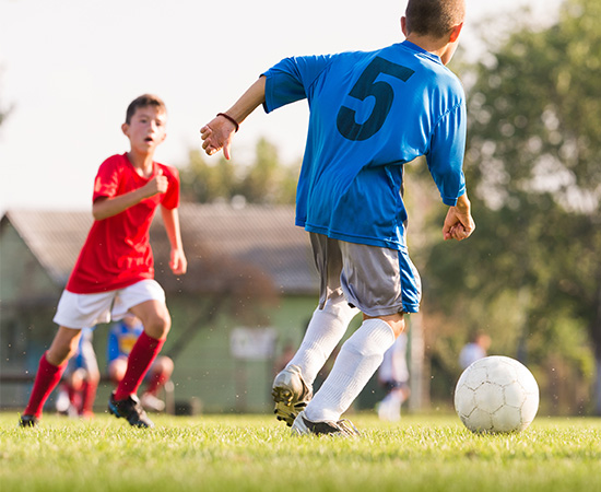 Two boys playing soccer on a grass field; one in a red jersey, the other in a blue jersey with the number 5.