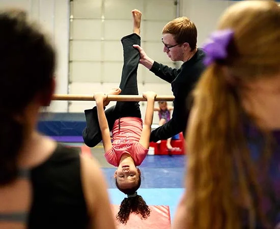 Ein junges Mädchen hängt kopfüber an einem Turnbalken und lächelt, während ein Ausbilder sie stützt. Zwei andere Kinder mit Pferdeschwänzen schauen im Vordergrund zu. Sie tragen personalisierte Fußballtrikots in einer mit Matten und Geräten gefüllten Turnhalle.
