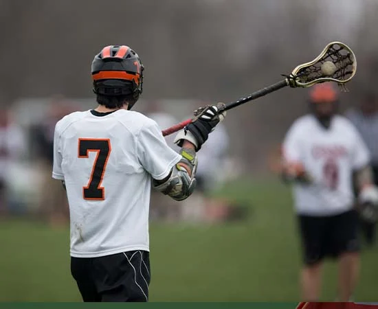 Ein Lacrosse-Spieler in einem weißen Trikot mit der Nummer 7, der einen Helm trägt, hält einen Lacrosse-Schläger in der Mitte des Spielfelds - so wie die Spieler ihr Aussehen mit personalisierten Fußballtrikots eines Top-Designers gestalten.