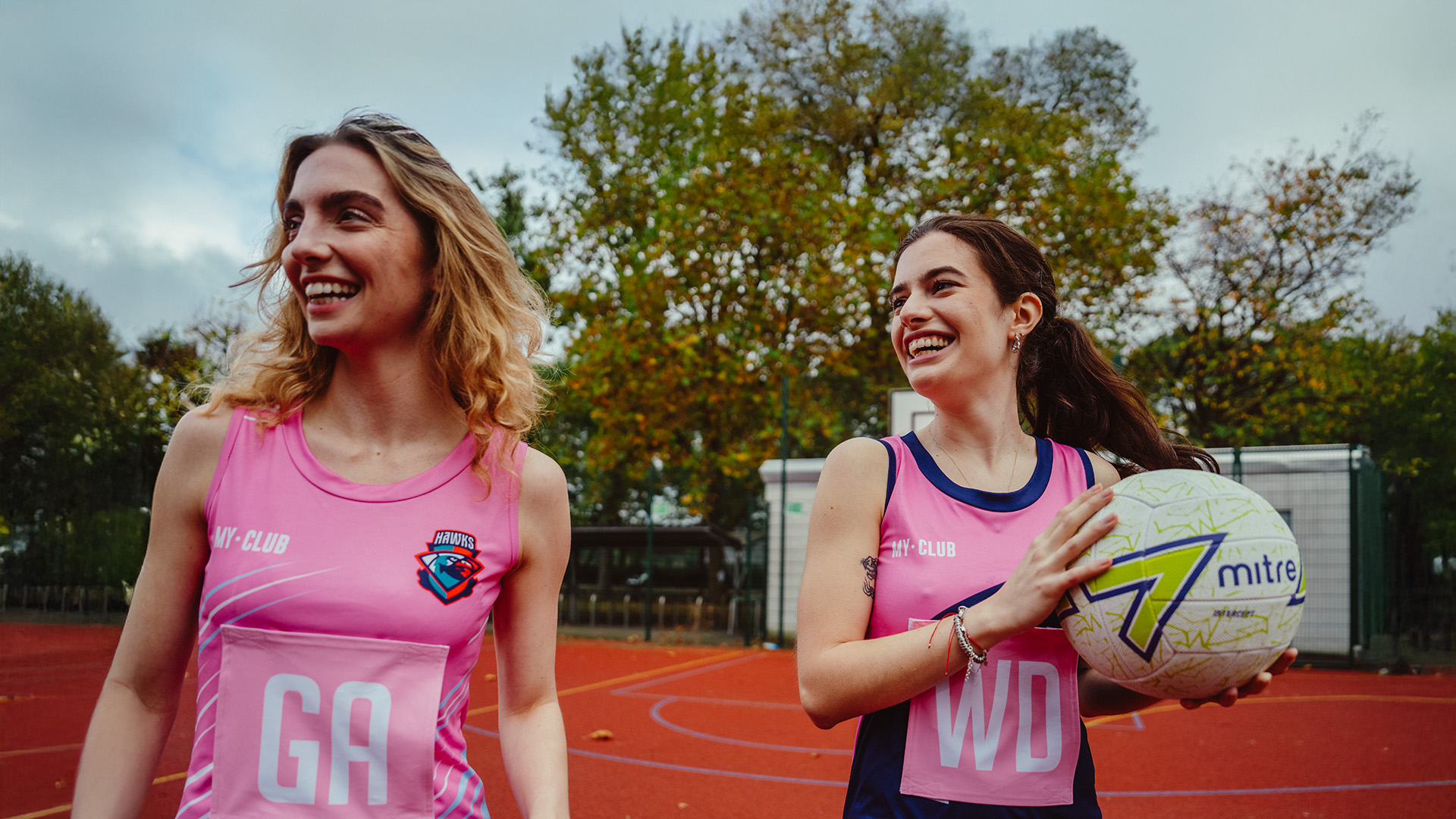 Two young women in bespoke sportswear smile and walk on an outdoor court. One holds a netball. Trees and a sports fence are visible in the background on a cloudy day.
