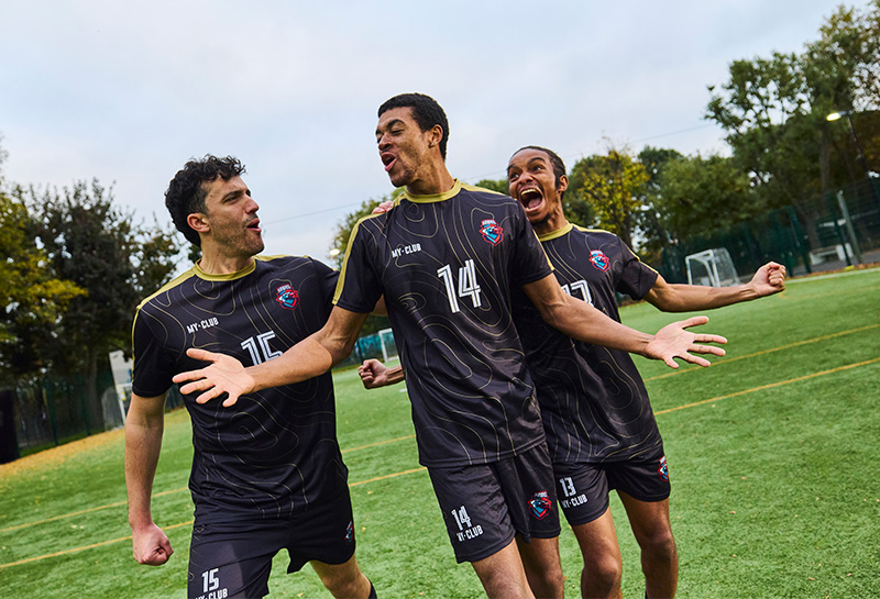 Three soccer players in matching jerseys celebrate on a sports field with trees in the background.
