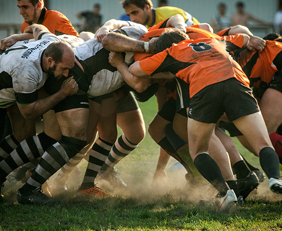 Two rugby teams engage in a scrum on a grassy field, with players in orange and black uniforms pushing against those in white and black. Dust is visible at their feet.