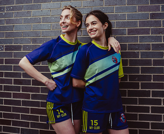 Two people in matching blue sports uniforms with green accents stand against a brick wall, smiling and posing together.