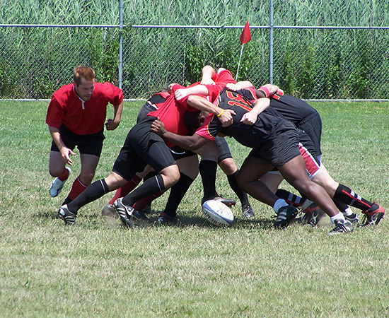 Rugby players in red and black jerseys engage in a scrum on a grassy field with a chain-link fence and tall grass in the background.