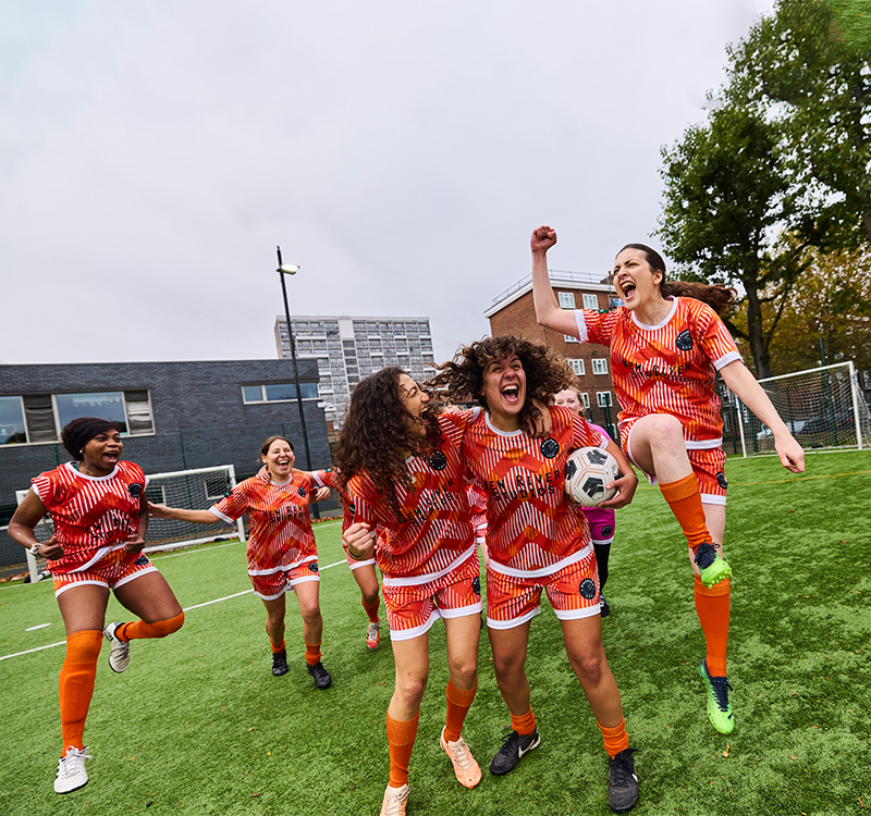 Un groupe de femmes en uniforme de football orange fait la fête sur un terrain, avec une joueuse tenant un ballon de football et d'autres personnes qui l'encouragent.