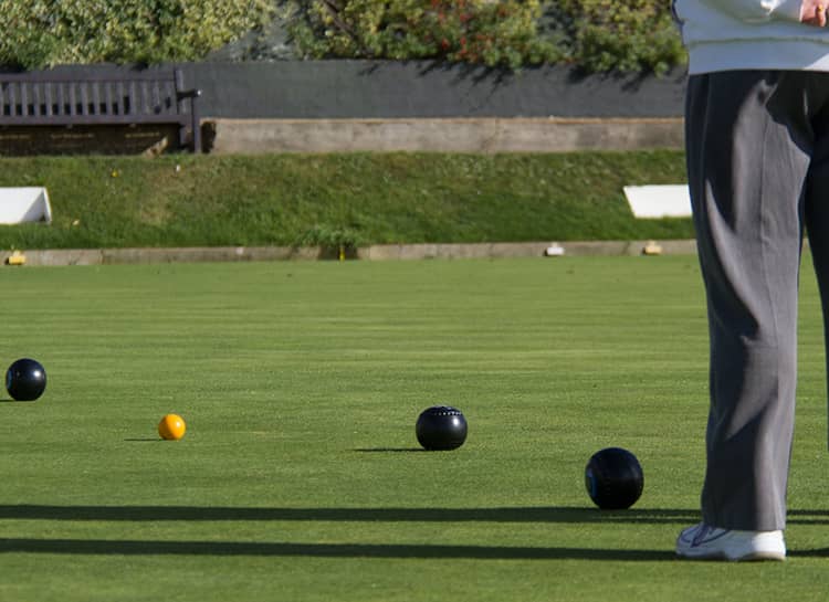 Una persona está de pie en un cuidado campo de bolos sobre césped, con varios bolos negros y una pequeña bola amarilla esparcidos cerca. Al fondo se ve un banco de madera y vegetación.