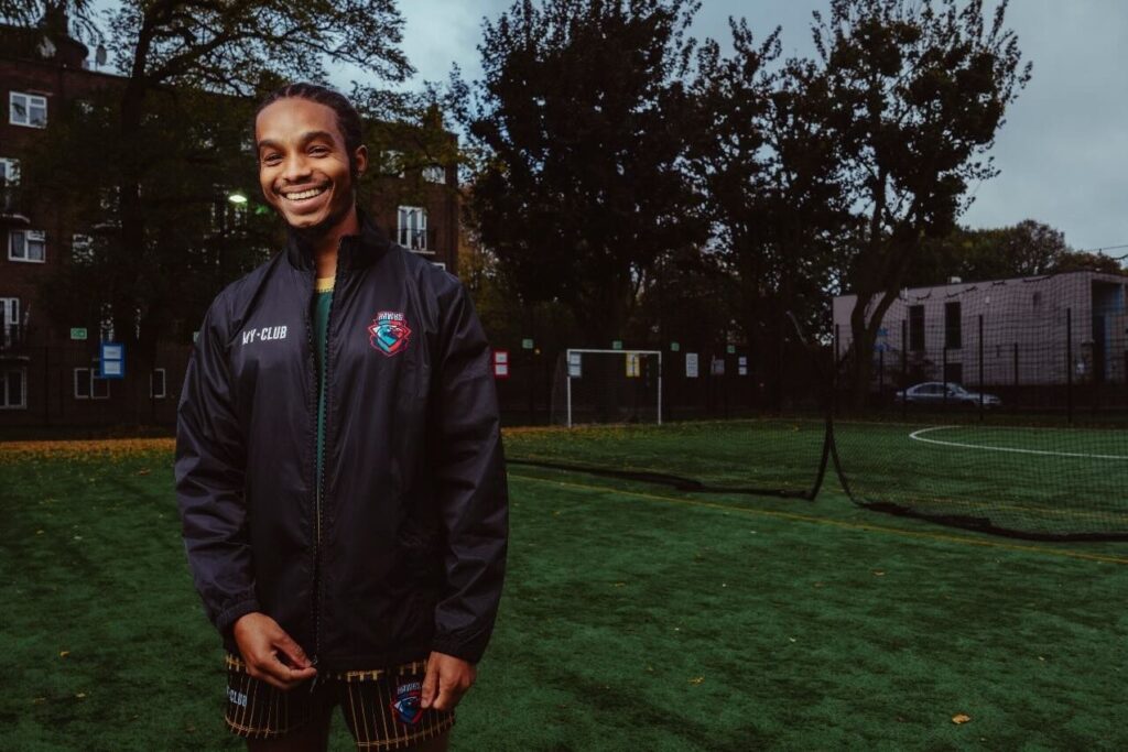 Un homme souriant portant une veste de sport noire se tient sur un terrain de football vert à la tombée de la nuit, où les joueurs montrent souvent leurs tenues de football personnalisées. Des arbres, un poteau de but et des bâtiments sont visibles à l'arrière-plan.