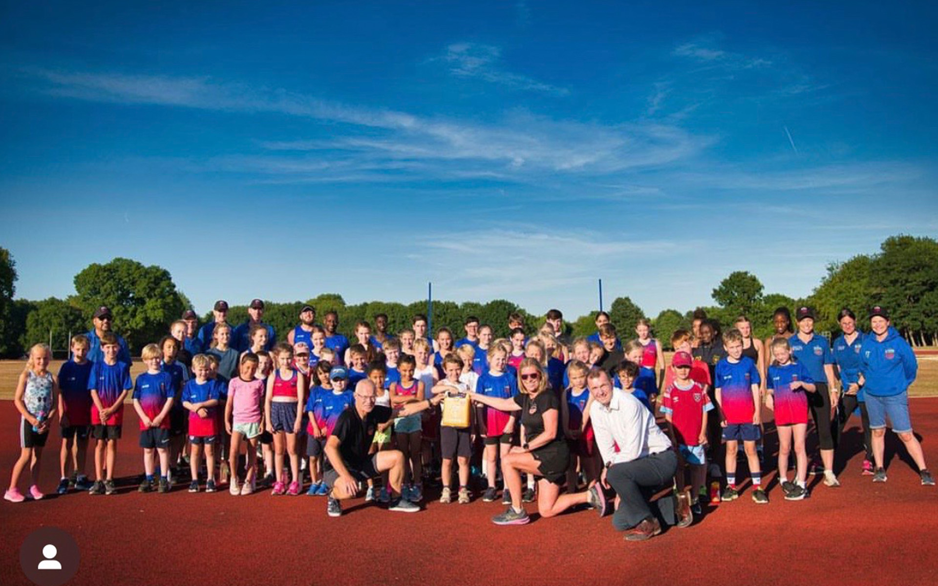 A large group of children and adults pose together on an outdoor running track under a clear blue sky. Most are wearing sports clothing, and some adults kneel at the front. Trees line the background.