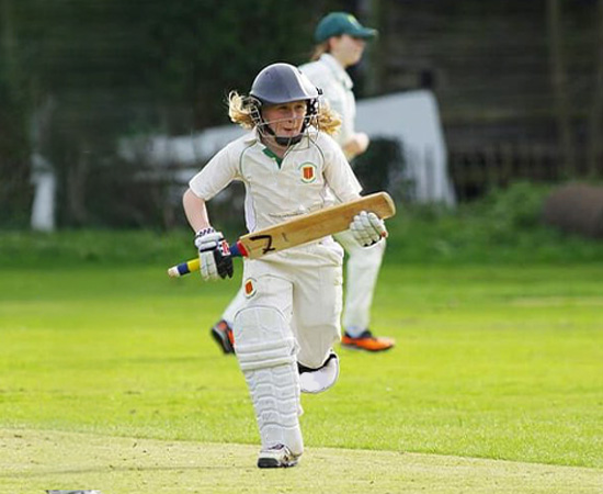 Youth cricket player in full gear running on a grassy field, holding a bat. Another player is in the background.