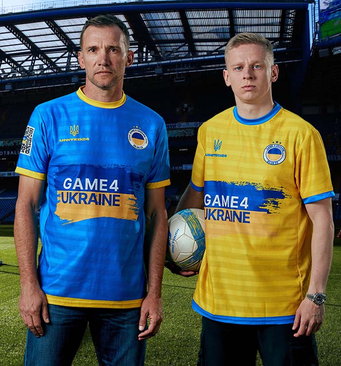 Dos hombres en un campo de fútbol con camisetas azules y amarillas de Game4Ukraine. Uno de ellos sostiene un balón. Al fondo se ven los asientos del estadio.