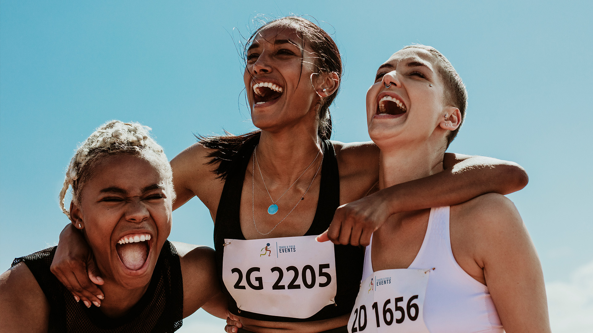 Drei Frauen in Sportkleidung mit Startnummern feiern fröhlich miteinander, lächeln und umarmen sich vor einem strahlend blauen Himmel.